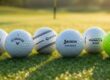 new year models premium golf balls, specifically a Titleist Pro V1, Callaway Chrome Tour, TaylorMade TP5, Srixon AD333, Pinnacle Rush, TopFlite XL resting on a pristine, dew-covered green at sunrise.