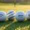 new year models premium golf balls, specifically a Titleist Pro V1, Callaway Chrome Tour, TaylorMade TP5, Srixon AD333, Pinnacle Rush, TopFlite XL resting on a pristine, dew-covered green at sunrise.